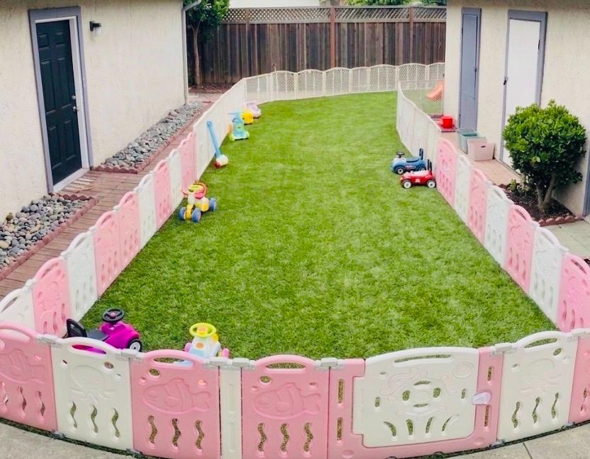Large fenced outdoor play yard with soft turf and ride-on toys