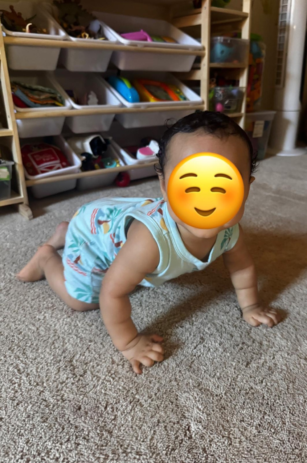 Infant crawling on the carpet near organized toy shelves with face covered for privacy