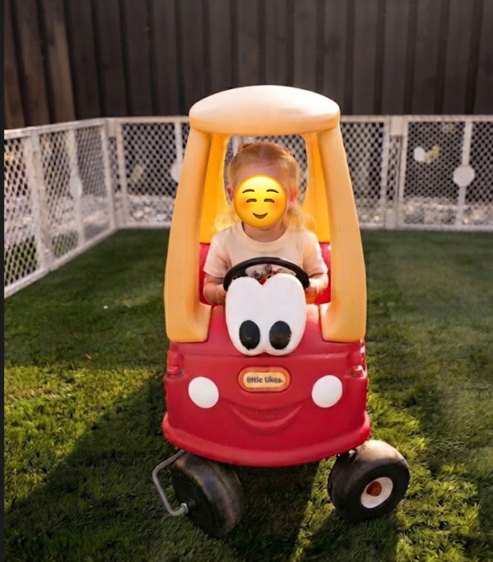 Child playing in a ride-on car in the fenced outdoor yard with face covered for privacy