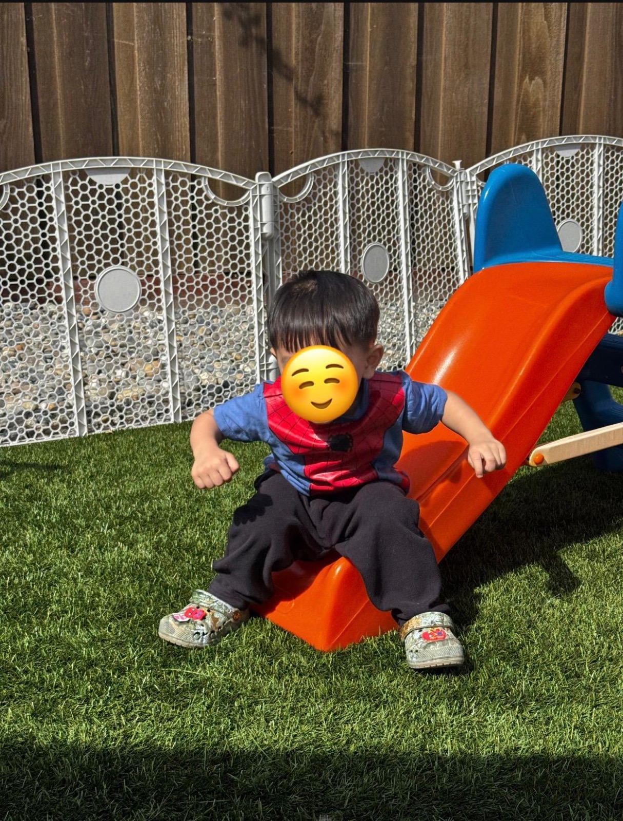Child sitting on a small slide in the fenced outdoor play yard at Ale's Home Childcare with face covered for privacy
