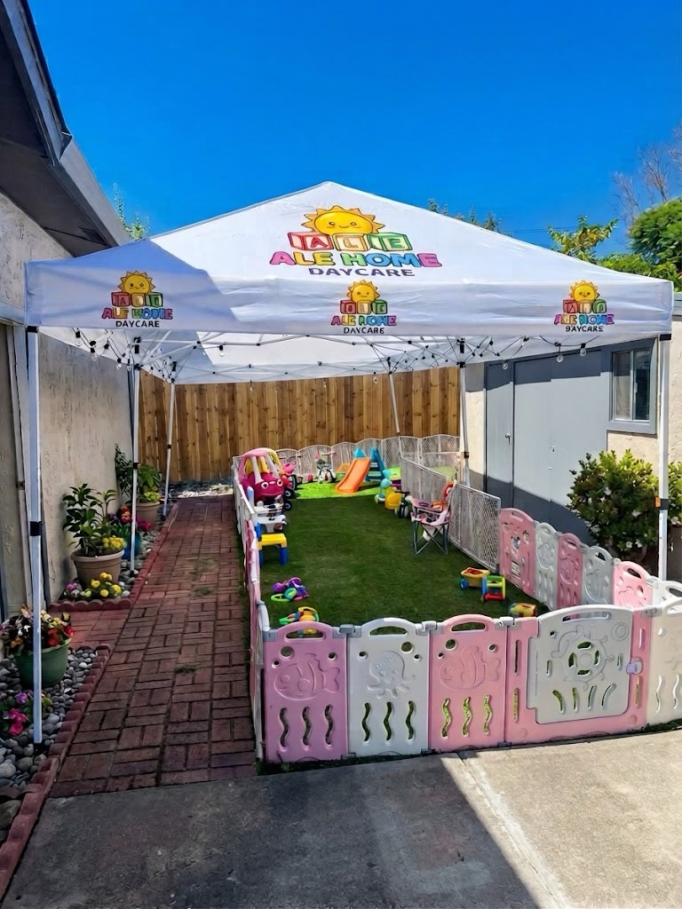 Large fenced outdoor play yard with soft turf, ride-on toys, and Ale Home Daycare canopy for shade