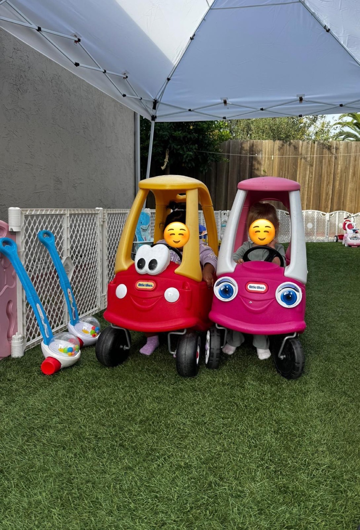 Children playing with ride-on cars in the shaded fenced outdoor yard with faces covered for privacy